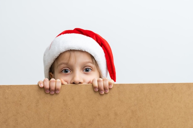 크리스마스 모자와 함께 웃는 어린 소녀의 초상화입니다. 빈 휴일 보드 뒤에 엿보는 아이. 흰색 배경에 고립 된 크래프트 종이 패널 뒤에 포즈를 취하는 행복 한 귀여운 아이. 프리미엄 사진