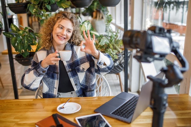 Бесплатное фото Портрет улыбающейся женщины-блогера в повседневной одежде, позирующей перед камерой с чашкой кофе