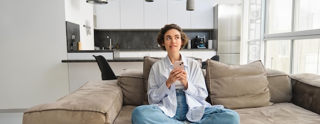 Бесплатное фото Портрет улыбающейся женщины, общающейся в приложении для смартфонов, сидящей дома на диване и использующей мобильный телефон