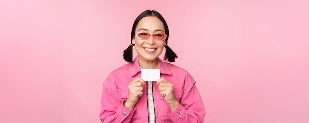 スタイリッシュな現代アジアの女の子の肖像画は割引クレジットカードを示し、ピンクの背景のコピースペースの上に立ってショッピングの非接触型の概念を支払うことを喜んで見える