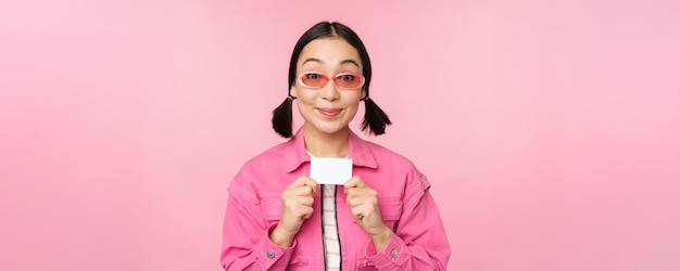 Портрет стильной современной азиатской девушки показывает дисконтную кредитную карту и выглядит довольным, оплачивая бесконтактную концепцию покупок, стоящую на розовом фоне
