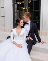 Бесплатное фото Портрет нежной супружеской пары жениха, целующего в лоб жену в красивом белом платье влюбленная пара, сидящая на лестнице и наслаждающаяся церемонией в день свадьбы