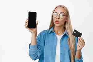 Бесплатное фото Портрет женщины в очках с любопытством показывает интересное приложение на мобильном телефоне и концепцию кредитной карты онлайн-покупок и бесконтактных платежей на белом фоне