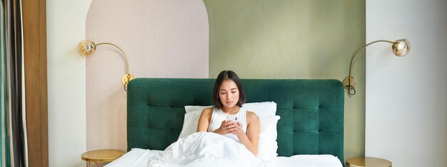 無料写真 羽毛布団の下のベッドに横たわり、深刻な顔でメッセージを読んでスマートフォンを見ている女性のポートレート