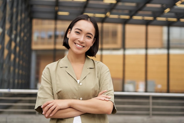 無料写真 自信を持って立っているアジアの若い女性の肖像画は、胸に腕を交差させ、屋外でポーズをとって微笑んでいます