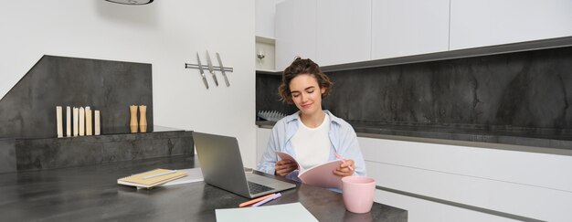 Бесплатное фото Портрет молодой красивой женщины учится дома, переворачивает свою тетрадь, работает из дома, использует ноутбук
