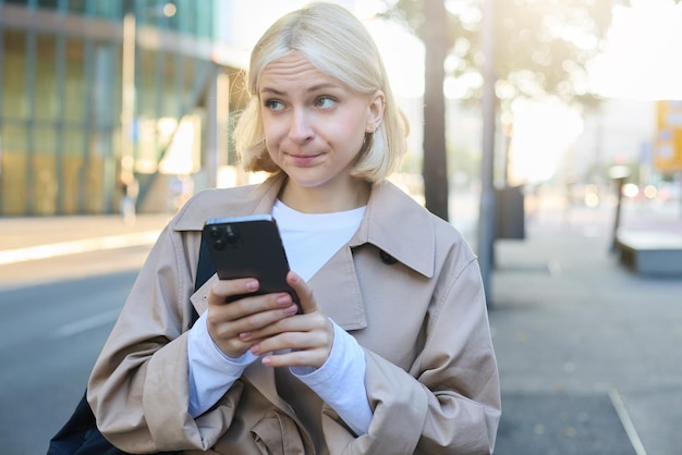 Бесплатное фото Портрет молодой блондинки, стоящей на улице, с неуверенным сомнительным выражением лица с помощью мобильного телефона
