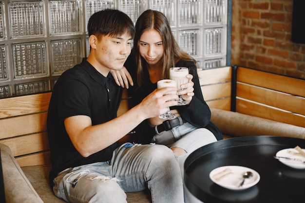 無料写真 カプチーノを手にレストランに座っている若いカップルの肖像画。黒髪の白人の女の子と黒髪のアジアの男の子を抱き締める