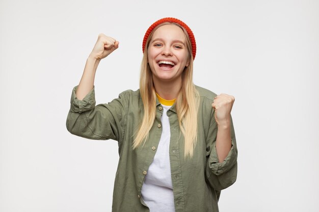 Бесплатное фото Портрет молодой длинноволосой блондинки, счастливо поднимающей кулаки и радостно смотрящей, в красной шляпе, оливковой рубашке и белой футболке, стоя на синем