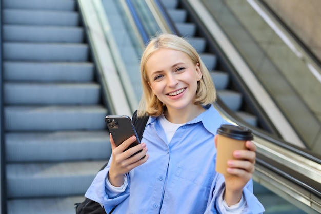 Бесплатное фото Портрет молодой современной женщины с рюкзаком смартфона, пьющей чашку кофе, стоящей рядом