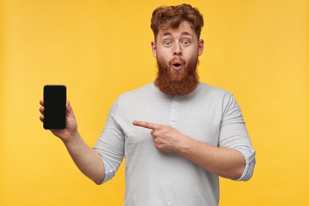 Портрет молодого позитивного рыжего мужчины с большой бородой, указывающего пальцем на дисплей своего телефона с пустой черной копией пространства
