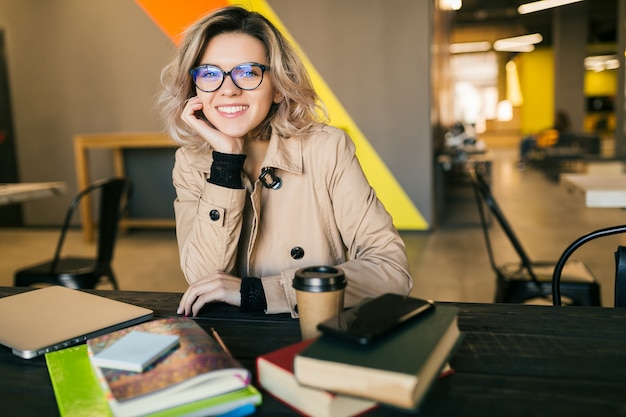 Бесплатное фото Портрет молодой красивой женщины, сидя за столом в тренче, работает на ноутбуке в офисе совместной работы, в очках, улыбаясь, счастливый, позитивный, на рабочем месте