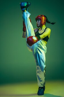 ネオン​の​光​の​緑​の​スタジオ​の​背景​の​上に​分離された​特別な​制服​で​キックボクシング​を​訓練する​若い​スポーティーな​女の子​の​肖像画