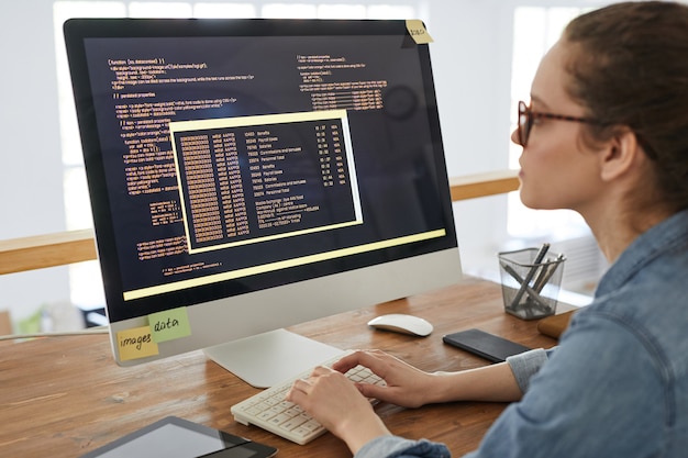 Портрет молодой женщины, пишущей программный код на экране компьютера, работая за столом в интерьере современного офиса, копией пространства