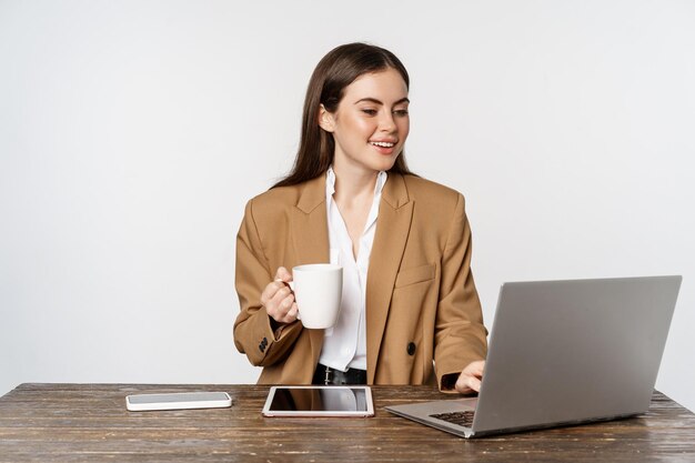 Portrait of office woman, ceo entrepreneur working on laptop, drinking coffee and answer clients on website, standing over white background