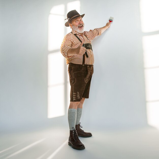 Portrait of Oktoberfest senior man in hat, wearing the traditional Bavarian clothes. Male full-length shot at studio on white background. The celebration, holidays, festival concept. Painting a wall.