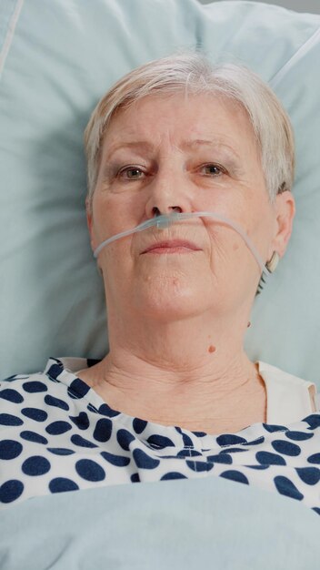 Portrait of old patient with disease having nasal oxygen tube while laying in bed. Close up of senior woman looking at camera and receiving healthcare treatment in hospital ward.