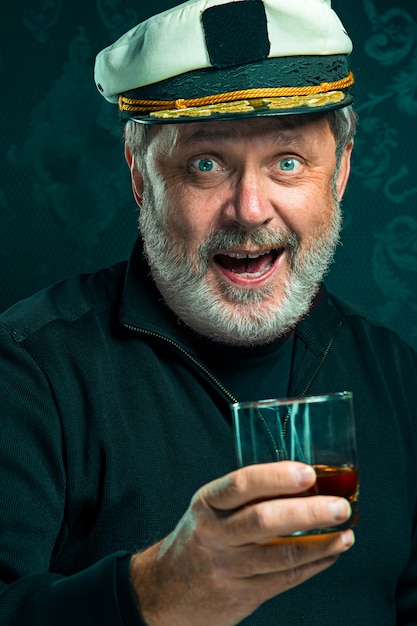 Портрет старика в роли капитана в черном свитере и шляпа, пьющая коньяк на черной студии