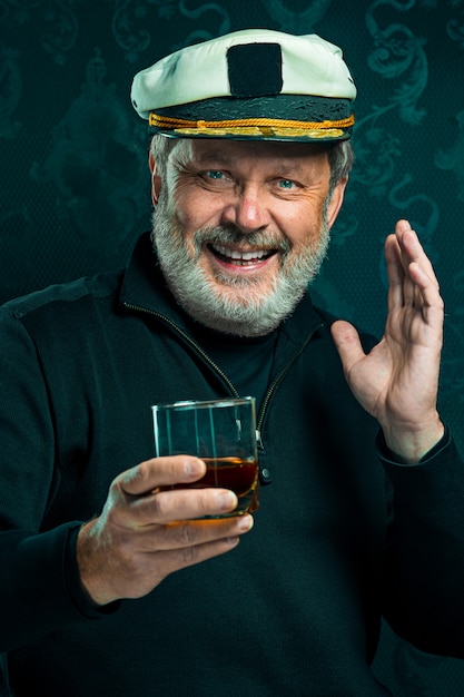 Portrait of old sailor man as captain in black sweater and hat drinking cognac on black studio