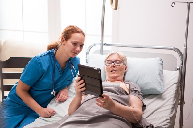 タブレットコンピュータを使用してナーシングホームで老婆の肖像画。引退した女性と女性看護師。