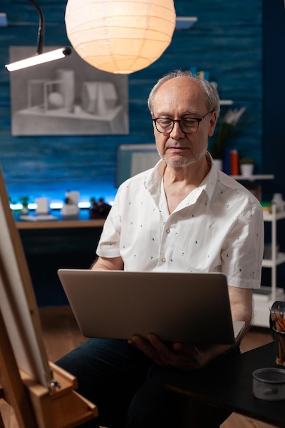 Portrait of older man looking to sell artwork on internet using portable computer in home studio. Senior artist spending evening sitting in front of easel holding laptop looking at online art gallery.