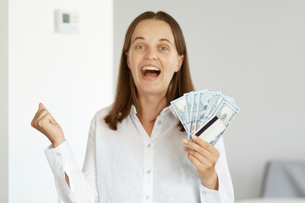 Portrait of optimistic extremely happy woman wearing white shirt, standing indoor in light room with credit card and money in hands, winning lottery, clenching fist and screaming happily. Premium Photo