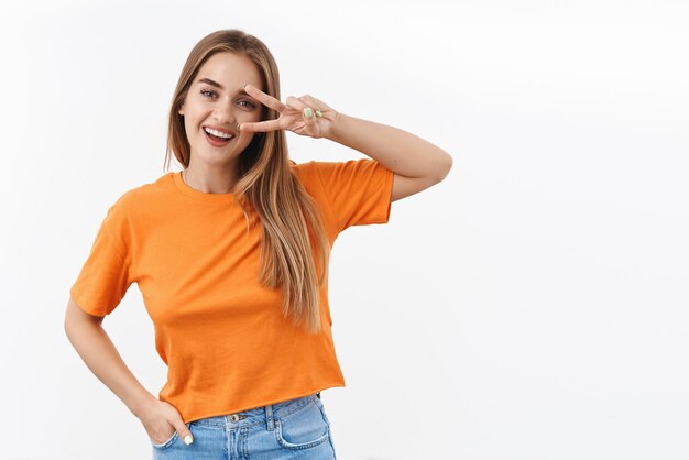 주황색 티셔츠를 입은 낙천적인 행복한 금발 소녀의 초상화, 눈 위에 평화 표시를 보여주고 웃고