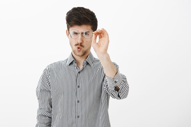 Portrait of ordinary adult european male student, taking off glasses and cleaning glass of eyewear, staring at goggles focused, folding lips to blow, standing over gray wall