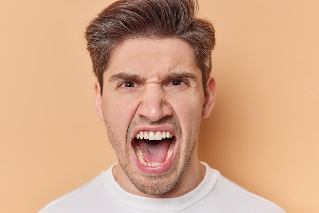 Portrait of outraged angry man keeps mouth opened screams with annoyance has aggressive expression poses against brown background. frustrated male model shouts loudly feels mad and irritated