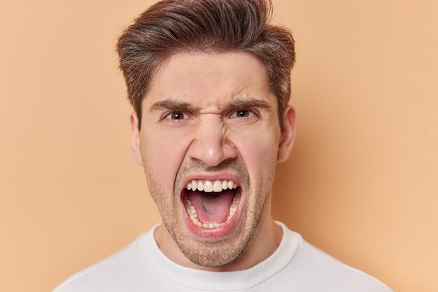 Portrait of outraged angry man keeps mouth opened screams with annoyance has aggressive expression poses against brown background. Frustrated male model shouts loudly feels mad and irritated