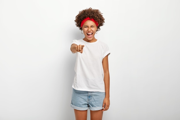 Портрет обрадованной афро-женщины смеется над чем-то забавным, указывает прямо в камеру, выражает хорошие эмоции, носит красную повязку на голову, футболку и шорты, модели над белой стеной.