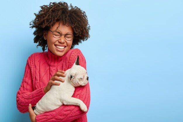 Portrait of overjoyed female dog owner holds small white puppy, laughs positively, being in good mood after outdoor walk with favourite pet, dressed in casaul jumper, has Afro hair. Animals concept