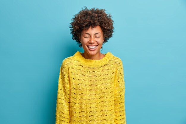 Portrait of overjoyed gentle woman smiles broadly closes eyes and shows white teeth wears casual yellow knitted jumper expresses positive emotions isolated over blue wall