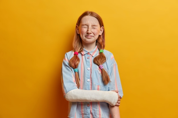 Portrait of overjoyed little girl cannot stop laughing, poses with closed eyes has ginger hair combed in pony tails dressed casually feels very happy has broken arm. Children, emotions, natural beauty