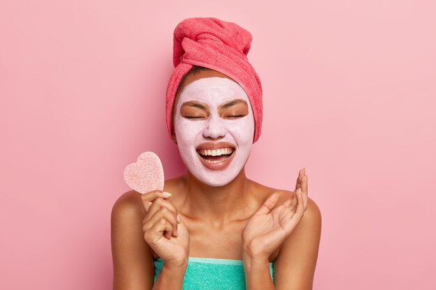 Portrait of overjoyed woman laughs happily, holds cosmetic sponge, raises palm being in high spirit, wears clay mask on face for removing wrinkles and clog pores, stands alone over pink wall