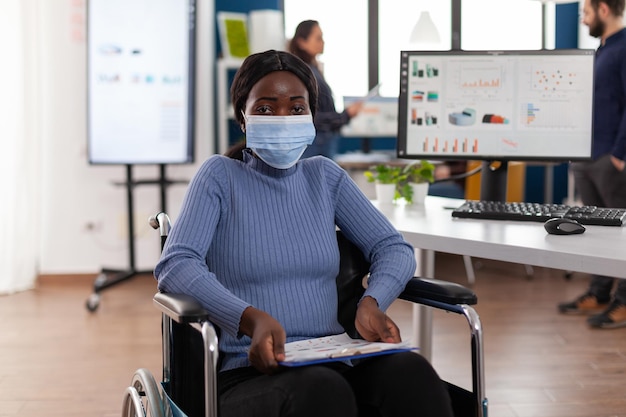 스타트업 회사 사무실에서 일하는 의료용 얼굴 마스크를 쓴 휠체어를 탄 마비된 아프리카계 미국인 여성 사업가의 초상화. 마케팅 전략을 분석하는 장애인 관리자