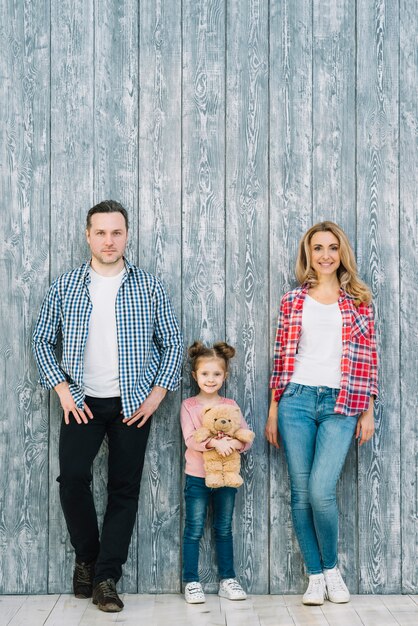 Portrait of parents standing with their daughter holding teddy bear against wooden background