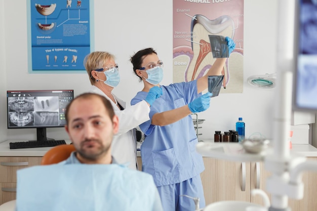 Portrait of patient with toothache waiting for medical procedure during stomatological consultation in dentistry office room. In background medical team analyzing teeth radiography. Medicine service