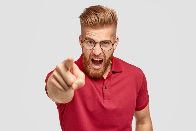 Portrait of peevish bearded guy with ginger trendy hairstyle, yells angrily at somebody, points with index finger directly at camera