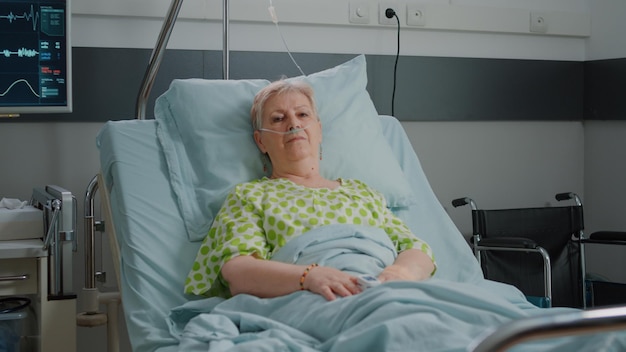 Portrait of pensioner waiting to receive medical support in hospital ward, laying in bed with nasal oxygen tube and IV drip bag. Aged patient preparing to cure sickness. Healthcare service