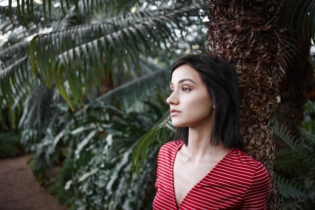 Portrait of pensive attractive young Caucasian woman in striped top spending day in jungle, standing at palm tree, looking ahead of her with thoughtul expression, deep in ideas, thoughts and dreams