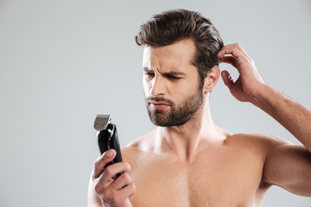 Portrait of a pensive bearded guy looking at electric shaver