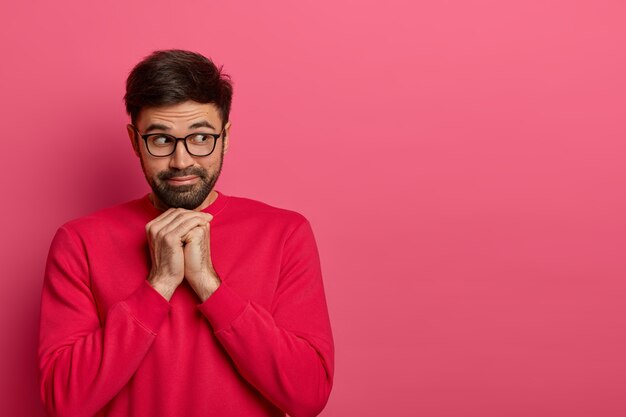 Portrait of pensive bearded man looks aside, keeps hands under chin, gazes with interest away, wears casual bright jumper and spectacles, thinks about something. Monochrome. Copy space aside for text