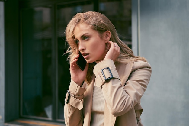 Ritratto di ragazza bionda pensosa in trench distoglie lo sguardo pensieroso parlando sul cellulare all'aperto