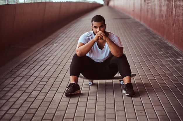 Portrait of a pensive dark-skinned guy dressed in a white shirt and sports shorts sitting on a skateboard on a footway under a bridge. Free Photo