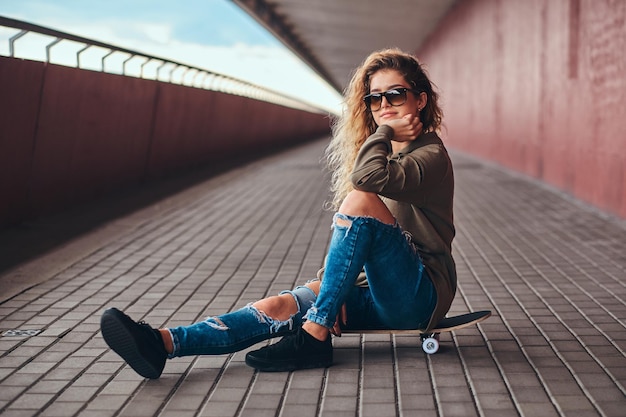 Portrait of a pensive girl in sunglasses dressed in a hoodie and ripped jeans sitting on a skateboard at a bridge footway.