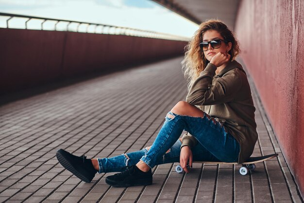 Портрет задумчивой девушки в солнцезащитных очках, одетой в толстовку с капюшоном и рваные джинсы, сидящей на скейтборде у моста.
