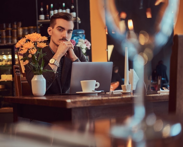 Portrait of a pensive handsome businessman with stylish beard and hair dressed in a black suit sitting with hand on the chin at a cafe with an open laptop, lookong at the camera.