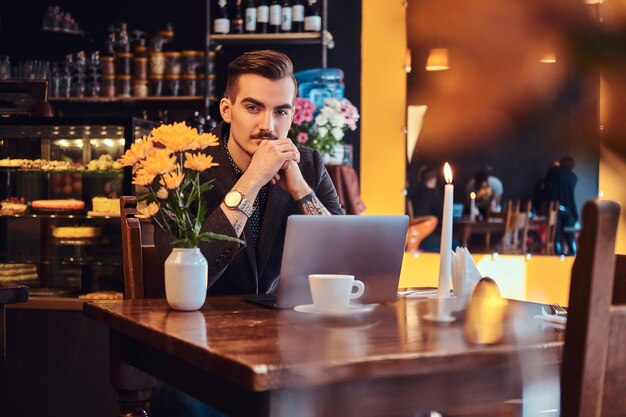 Портрет задумчивого красивого бизнесмена со стильной бородой и волосами, одетого в черный костюм, сидящего с рукой на подбородке в кафе с открытым ноутбуком и смотрящего в камеру.