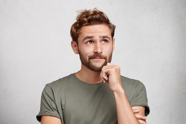 Portrait of pensive pleasant looking young male with beard and mustache, keeps hand under chin